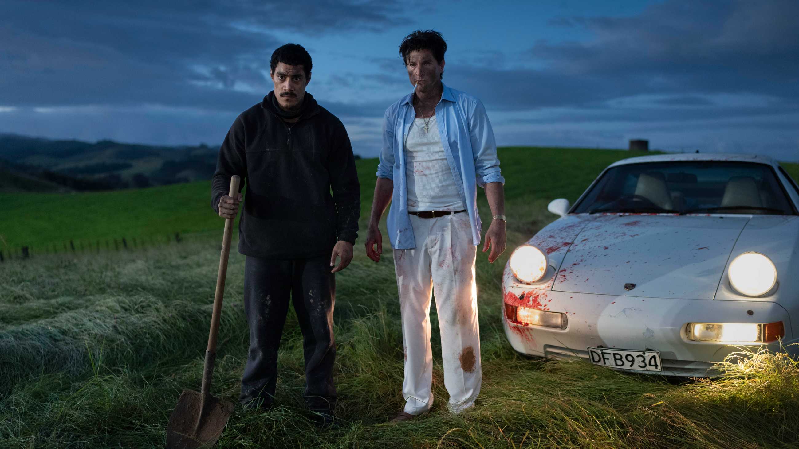 Mafu holding a shovel next to a dirtied Marco and a car with blood splatter in God Bless You, Mr. Kopu