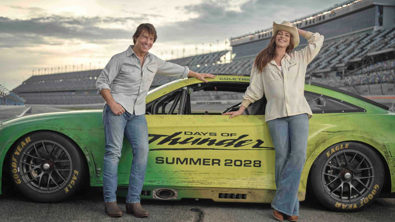 Tom Cruise and Anne Hathaway pose in front of a race car in Days of Thunder 2 promo.