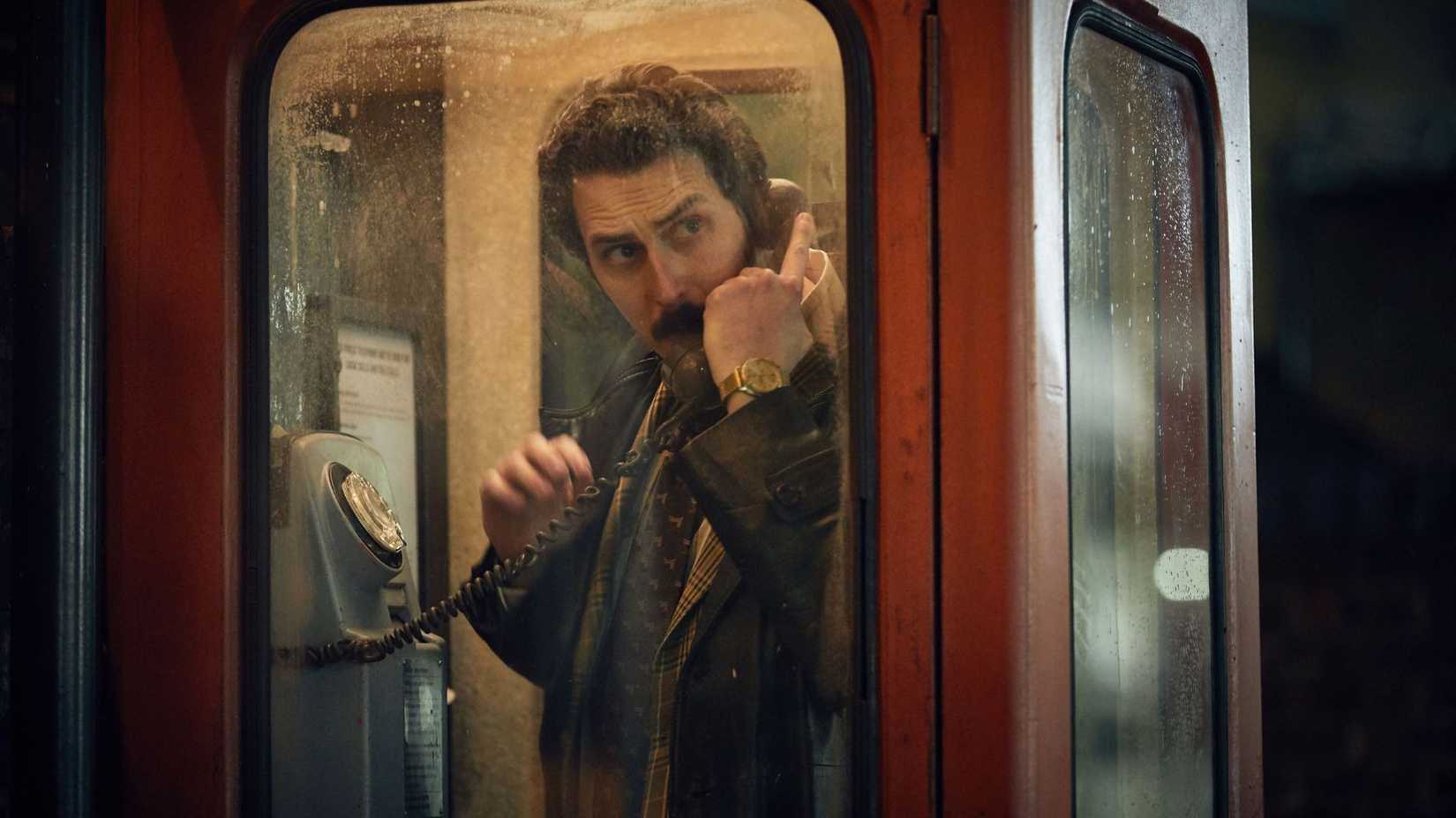 A white man with brown hair stands in an old-fashioned red British pay phone booth. He is looking over his shoulder at something.