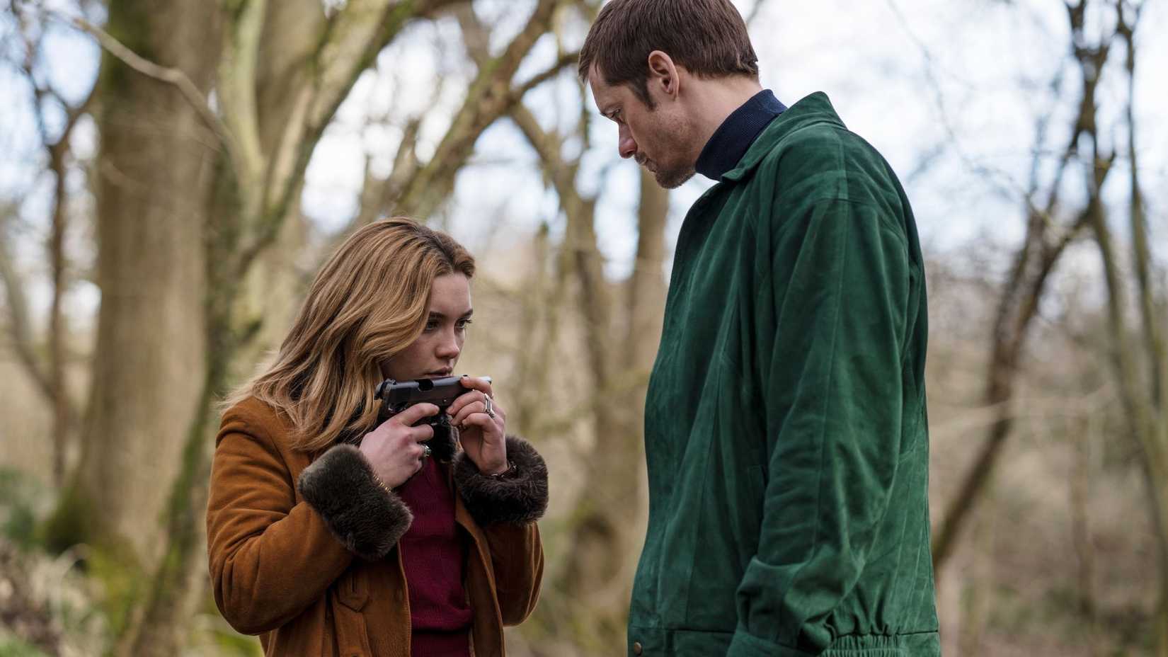 A very tall man in a green jacket and a very short woman in a brown coat stand in the woods. The woman is holding a gun.