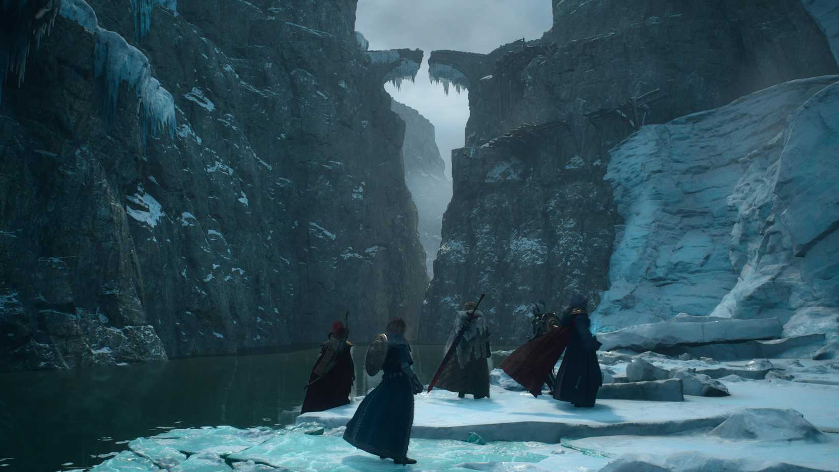 Five people stand on an icy river bank looking toward the cliffs above, where a broken bridge can be seen covered in icicles.