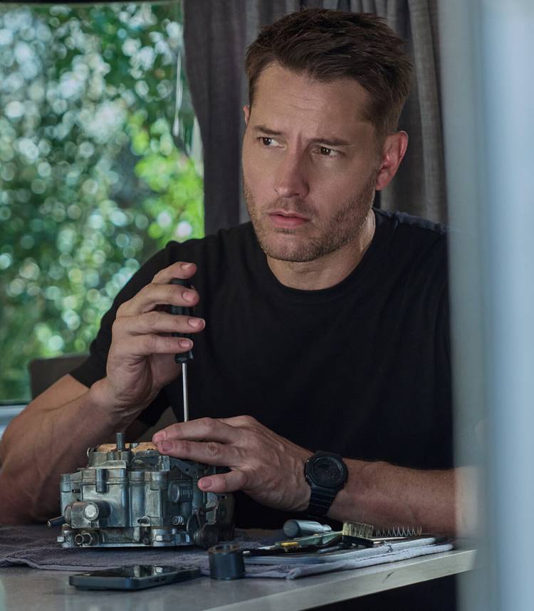 Justin Hartley's Colter looking in thought while working on an engine in his Airstream in Tracker season 4 vertical