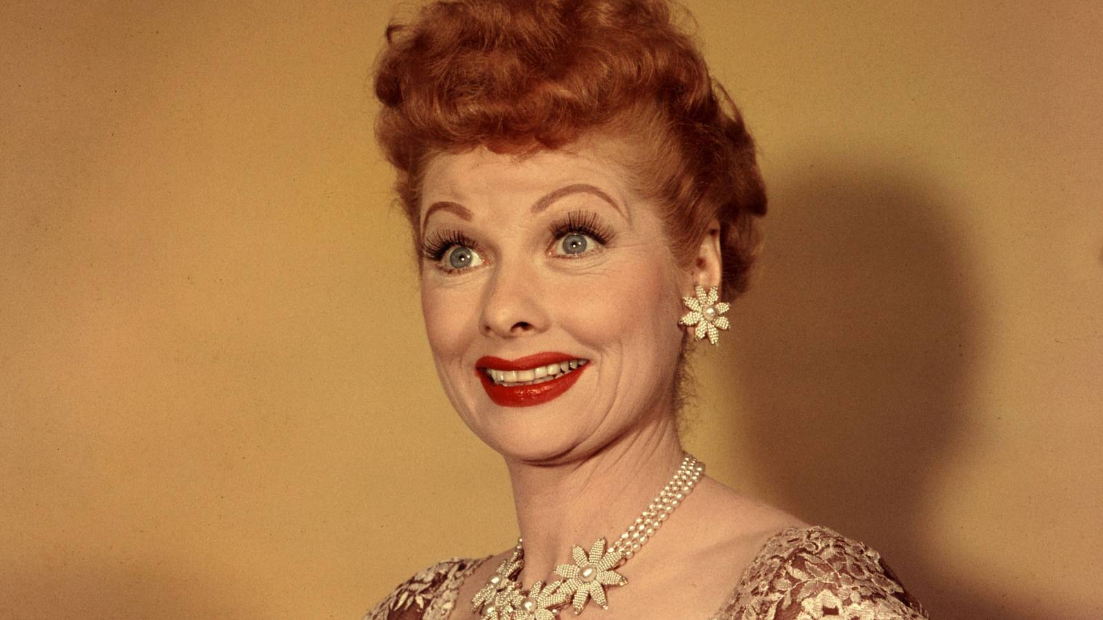 Lucy has red hair, red lipstick, and flower earrings and a necklace in the promo photo of I Love Lucy
