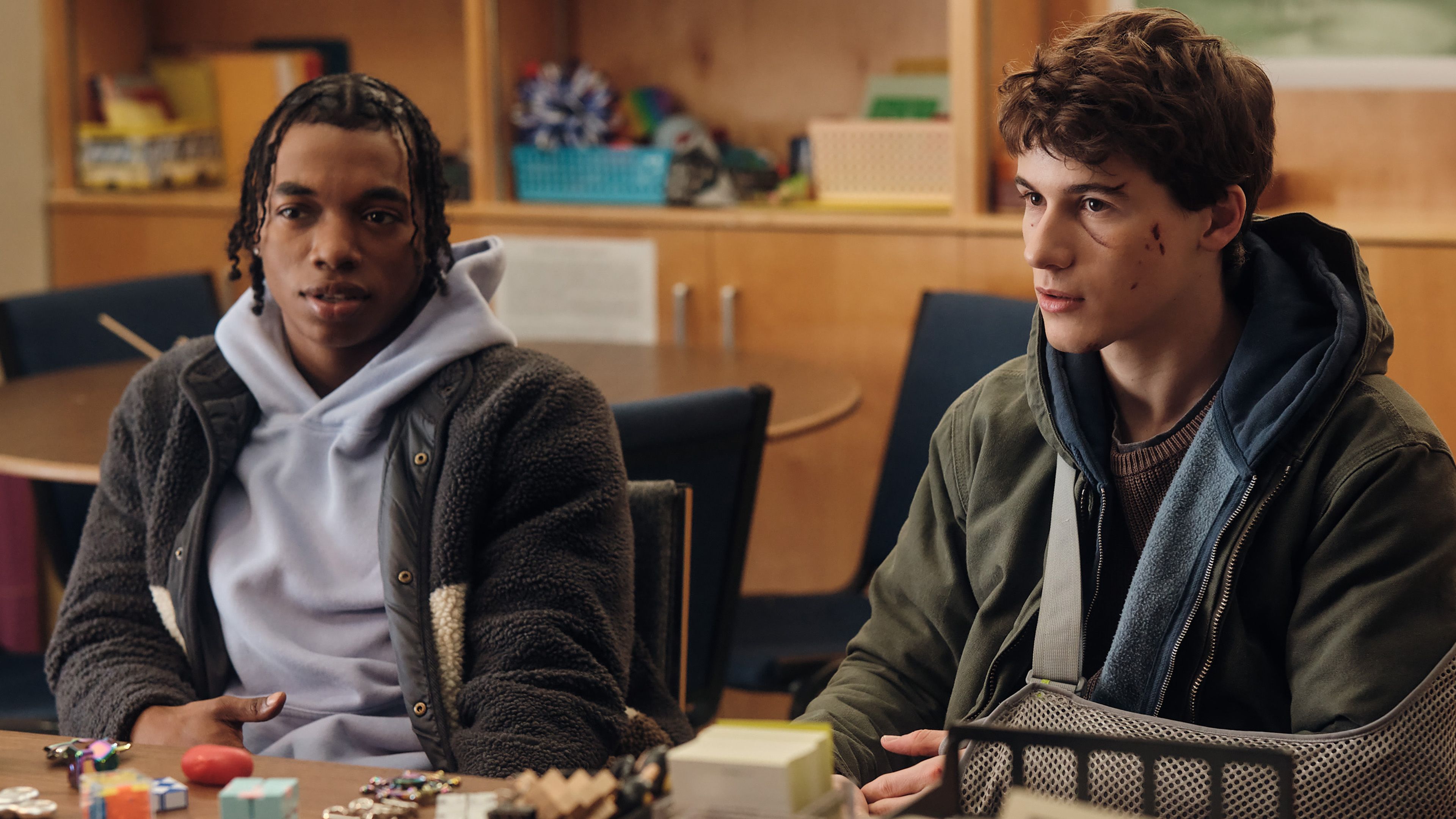 sam-nivola-s-sonny-and-maliq-johnson-s-vogel-looking-at-someone-while-sitting-down-next-to-each-other-in-phony Phony: Hulu’s New Mystery Series Is An Instantly Gripping & Enjoyable Ride | Review