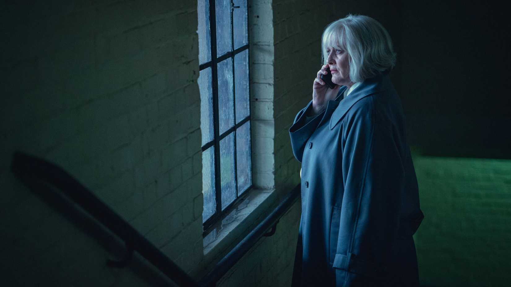 A woman with white hair stares out a window while talking on the phone.