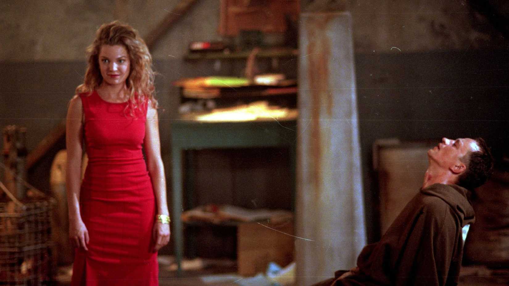 Glory wearing a red dress stands in front of a monk tied to a chair in Buffy